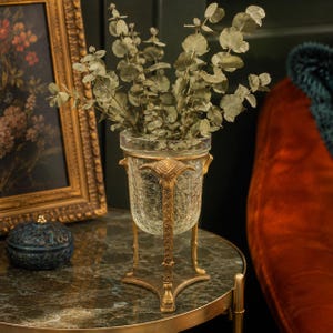 May include: A clear crackle glass vase held by a gold-toned metal stand featuring three legs and decorative ram head accents at the top. The vase contains a cluster of dried green eucalyptus stems with round leaves, creating a botanical display.