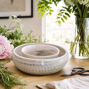 May include: Two ceramic bowls, one inside the other. The larger bowl is white with a textured pattern and pink accents. The smaller bowl has a pink and grey marbled design. The bowls are on a wooden table, with flowers and a vase in the background.