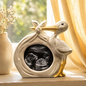 May include: A ceramic stork ornament carrying a baby in a bag. The stork is white with yellow beak and legs. The bag has a black oval with a baby ultrasound image.