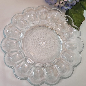 May include: Clear glass egg serving dish with a scalloped edge. The dish has 12 egg-shaped indentations around the rim and a textured centre. The glass is transparent, allowing light to pass through.