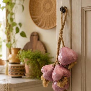 May include: A decorative hanging ornament featuring four glossy, light purple garlic bulbs attached to a thick, braided jute rope. The bulbs have textured, tan-coloured fibrous roots at the base and are suspended from a dark metal knob on a white cabinet.