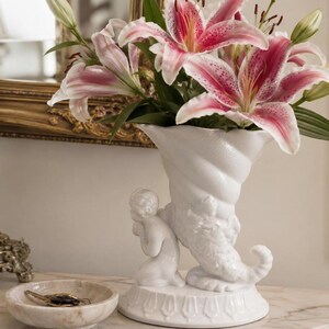 May include: A white ceramic cornucopia vase featuring a sculpted kneeling figure at the base, filled with pink and white lilies. Beside the vase sits a small, round, beige marble bowl containing a set of metal keys on a ring.