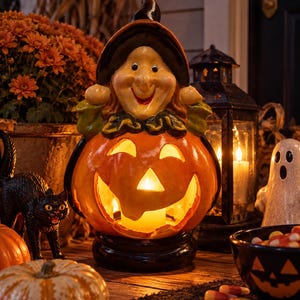 May include: A ceramic Halloween decoration featuring a smiling witch figure wearing a black hat, perched atop a glowing orange jack-o'-lantern with carved triangular eyes and a wide mouth, all set on a round black base with an internal light source.