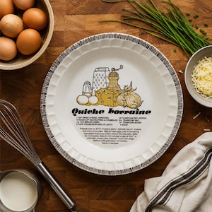 May include: A white ceramic pie dish with a fluted edge and black rim detail. The centre features a yellow and black illustration of ingredients and the text Quiche Lorraine, followed by a list of ingredients and baking instructions.