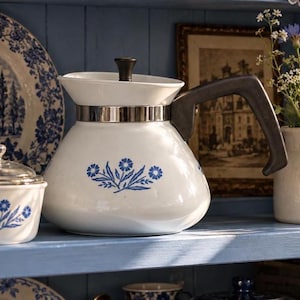 May include: A white ceramic teapot featuring a blue floral pattern on the front, a black handle, and a black lid knob. The teapot has a wide, rounded base and a metallic silver band encircling the neck, connecting the body to the lid assembly.