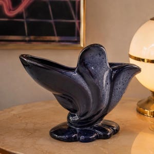 May include: A ceramic vase featuring a deep navy blue and mottled grey glaze. The vessel has an asymmetrical, organic shape with a wide, flared opening and a curved, petal-like structure that tapers down to a solid, rounded base.