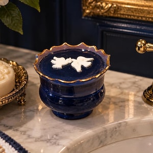 Peut inclure: Un pot en céramique bleu profond avec un couvercle festonné bordé d'or, orné de deux colombes blanches en relief. Le récipient possède un corps arrondi et une base sur pied, offrant une finition lisse et brillante avec des détails en relief.