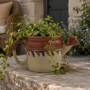 Peut inclure: Un arrosoir en céramique servant de pot de fleurs avec un bord brun brillant et une base crème tachetée. Le corps présente un motif peint à la main de traits diagonaux alternant brun et noir. Une plante verte retombante pousse à l'intérieur.