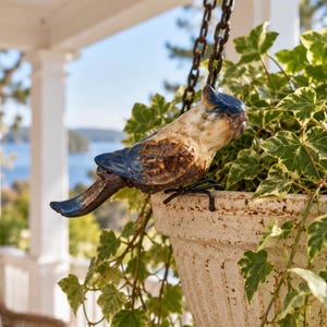 May include: A ceramic bird figurine with a glossy blue and cream glaze, perched on the rim of a weathered, off-white textured planter. The bird is attached to a dark metal chain, and the planter is filled with green and white variegated ivy.