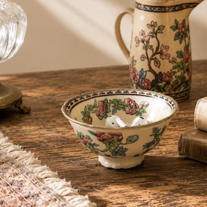 May include: A small, cream-coloured ceramic bowl featuring a hand-painted floral pattern with pink blossoms and green leaves, accented by a dark geometric border along the rim. The bowl contains two white wireless earbuds inside its basin.