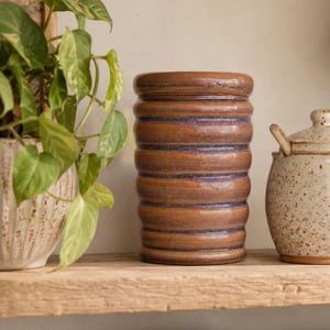 Peut inclure: Un pot en céramique cylindrique avec une glaçure brune brillante et une texture nervurée horizontale présentant de subtils accents bleus entre les rainures. Le récipient possède un couvercle assorti et repose sur une surface plane.