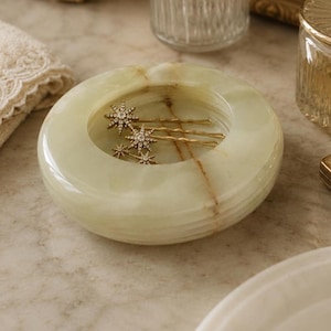 Peut inclure: Un plat rond en pierre vert pâle avec des veines brunes naturelles, contenant trois épingles à cheveux dorées. Chaque épingle présente un ornement en forme d'étoile incrusté de petites pierres scintillantes, reposant dans le bol poli et lisse.
