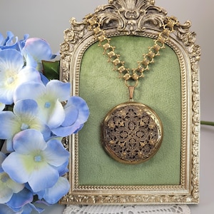 Peut inclure: Un collier locket doré de style vintage avec des détails de filigrane complexes. Le pendentif rond est suspendu à une chaîne décorative et est présenté sur un fond vert dans un cadre doré. Des fleurs d'hortensia bleues sont au premier plan.