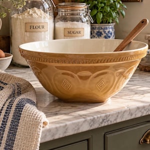 May include: A large, tan ceramic mixing bowl with a decorative embossed pattern around the rim and sides, featuring a smooth interior and a wooden spoon resting inside. The bowl has a glossy finish and a wide, circular opening for mixing ingredients.