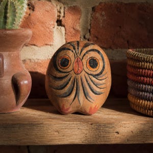 May include: A rounded, tan-coloured ceramic owl figurine featuring hand-painted black and orange details. The owl has large, circular eyes with orange rings, a small orange beak, and black feather-like markings across its body, with small orange feet.