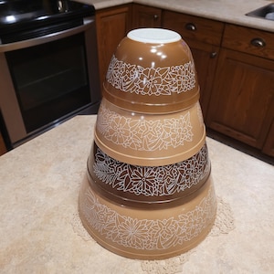 Puede incluir: Una pila de cuatro cuencos mezcladores vintage en tonos marrón, beige y blanco. Cada cuenco presenta un diseño floral blanco. Los cuencos están anidados, mostrando sus diferentes tamaños y formas. Utensilios de cocina.