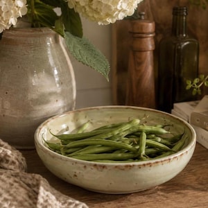 May include: A shallow, round, off-white ceramic bowl with a speckled finish and a rustic brown rim, filled with a pile of fresh green beans. The bowl has a smooth, slightly irregular texture and sits in a kitchen setting with other items.