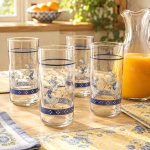May include: A set of four clear glass tumblers featuring a decorative pattern of white geese wearing blue polka-dot scarves. The design includes horizontal blue bands with white dots and small yellow accents, arranged around the cylindrical glasses.
