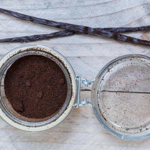 May include: An open glass jar filled with dark brown powder, next to an empty jar. Two dark brown vanilla pods are crossed above the jars. The jars have metal clasps and are on a light wooden surface.
