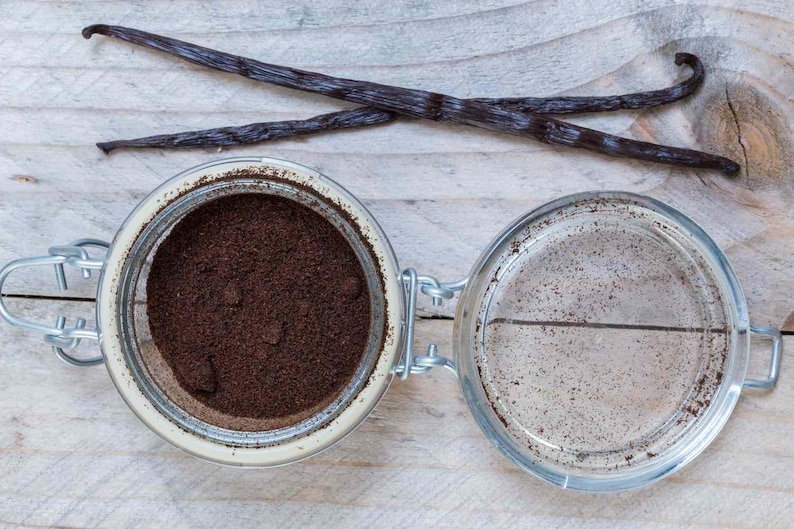 May include: An open glass jar filled with dark brown powder, next to an empty jar. Two dark brown vanilla pods are crossed above the jars. The jars have metal clasps and are on a light wooden surface.