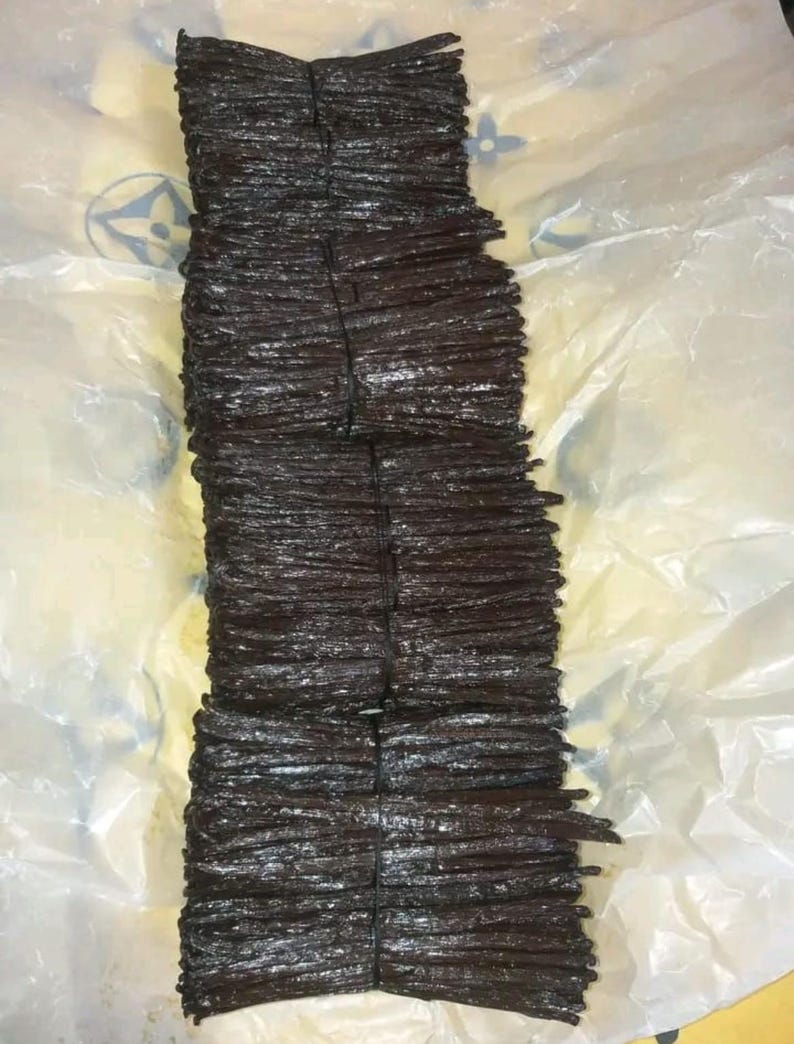 May include: Several bundles of dark brown vanilla pods tied with black twine. The pods are stacked on top of each other. The background is a white and yellow plastic sheet.