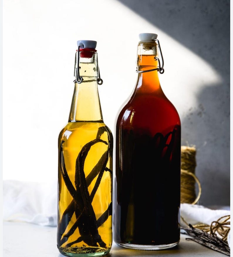 May include: Two glass bottles filled with liquid. One bottle contains a light yellow liquid with dark vanilla beans, and the other has a dark red liquid. Both bottles have swing-top closures.