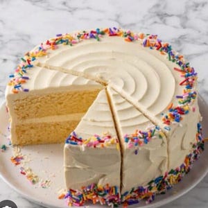 May include: A round cake with white icing and colourful sprinkles. A slice has been cut and removed, revealing the yellow cake layers. The cake sits on a white plate, ideal for a celebration.