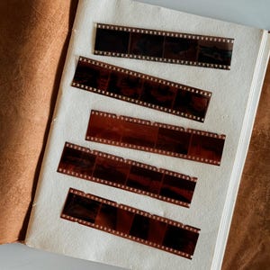 May include: An open vintage photo album with five strips of developed film. The film strips are brown and black, with visible frames and sprocket holes. The album has a light cream-colored page and a brown leather cover.