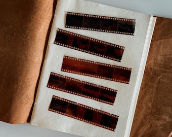 May include: An open vintage photo album with five strips of developed film. The film strips are brown and black, with visible frames and sprocket holes. The album has a light cream-colored page and a brown leather cover.