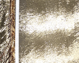 May include: Close-up of gold sequin fabric. The fabric is covered in small, shiny sequins that reflect light, creating a shimmering effect. A white ruler is placed vertically along the left side of the fabric, providing a measurement reference.