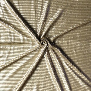May include: A close-up shot of a gold-colored fabric with a horizontal striped pattern. The fabric is folded and draped, creating shadows and highlights that emphasize the texture and sheen of the material. The fabric appears to be made of sequins or a similar reflective material.
