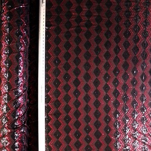 May include: A roll of black and red sequin fabric with a geometric diamond pattern. The fabric is covered in small, shiny sequins that catch the light. A ruler is placed vertically along the fabric.