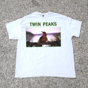 Può includere: T-shirt bianca con la scritta verde "TWIN PEAKS" sopra un'immagine incorniciata. L'immagine mostra un uccello marrone posato su un ramo, con uno sfondo sfocato di un lago e alberi.