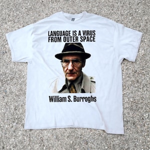 May include: White t-shirt with a portrait of William S. Burroughs, wearing a hat and glasses, and the text "LANGUAGE IS A VIRUS FROM OUTER SPACE" above his image, with his name below. The shirt is made of a soft material.