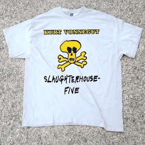 Könnte beinhalten: Weißes T-Shirt mit dem Text "KURT VONNEGUT" über einer gelben Totenkopf- und gekreuzten Knochen-Grafik. Darunter steht "SLAUGHTERHOUSE-FIVE". Das T-Shirt liegt flach auf einer strukturierten Oberfläche.