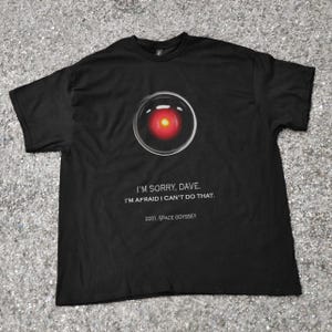 Peut inclure: T-shirt noir avec un motif graphique d'un design en forme d'œil rouge et blanc. Le texte "I'M SORRY, DAVE. I'M AFRAID I CAN'T DO THAT. 2001: SPACE ODYSSEY" est imprimé en dessous. Un tee-shirt sur le thème de la science-fiction.