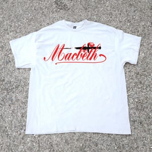 May include: White t-shirt with the word "Macbeth" in red script, a black sword, and a red rose design. The shirt is made of a soft material and has a crew neck.