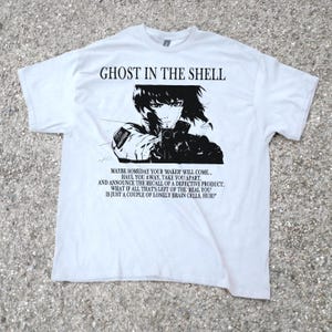 Può includere: T-shirt bianca con grafica in bianco e nero di "Ghost in the Shell". Il design include un personaggio anime con una pistola, con testo sopra e sotto l'immagine. Il testo recita "GHOST IN THE SHELL" e una citazione.