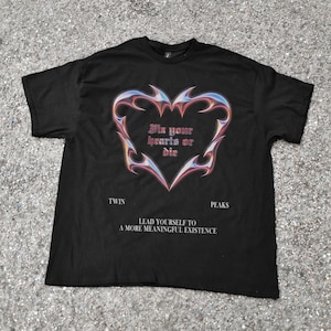May include: Black t-shirt featuring a heart-shaped design with flame-like edges in shades of blue, pink, and yellow. The text "Fix your hearts or die" is inside the heart. Below, the shirt reads "Twin Peaks" and "Lead yourself to a more meaningful existence."