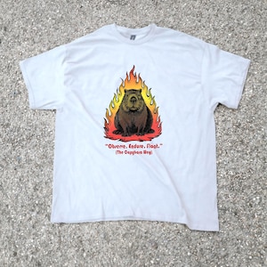 Può includere: T-shirt bianca con un castoro marrone circondato da fiamme. Il testo "Observe. Endure. Float." (The Capybara Way) è sotto l'immagine. La maglietta è realizzata in materiale morbido.