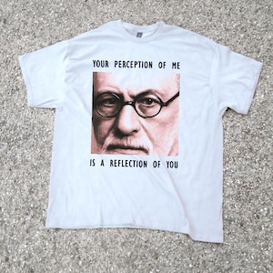 May include: White t-shirt featuring a portrait of a man wearing round glasses. The text "YOUR PERCEPTION OF ME IS A REFLECTION OF YOU" is printed above and below the portrait in black font. The shirt is laid out on a gray surface.