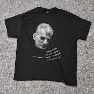 Puede incluir: Camiseta negra con un retrato en escala de grises de un hombre con gafas de sol. La camiseta incluye la cita "DANCE FIRST. THINK LATER. IT'S THE NATURAL ORDER. -SAMUEL BECKETT" en texto blanco. La camiseta es de tela suave.