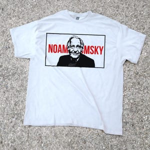 May include: White t-shirt featuring a black and red graphic of Noam Chomsky. The graphic is a portrait of Chomsky with the name "NOAM CHOMSKY" in red letters above the portrait. The t-shirt is made of a soft material.