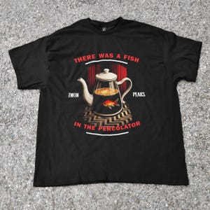 May include: Black t-shirt featuring a graphic design with the text "THERE WAS A FISH IN THE PERCOLATOR" and "TWIN PEAKS". The design includes a teapot with a fish inside, set against a red curtain backdrop.