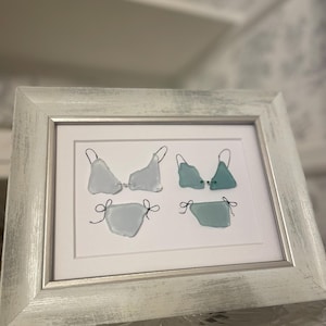 May include: A framed wall art piece featuring two bikini sets crafted from smooth sea glass pieces in shades of light blue and teal. The glass fragments are connected with thin black wire shaped into straps and bows, set against a white background.