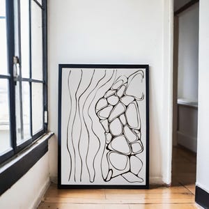May include: Black and white abstract art print in a black frame. The artwork features a design of organic shapes and lines. The print is displayed on a wooden floor next to a window.