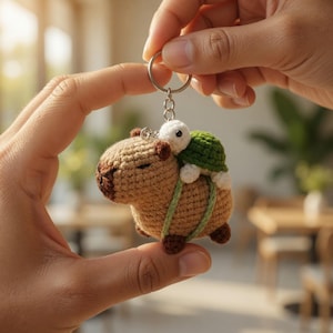 May include: A handmade crochet keychain featuring a brown capybara carrying a green-shelled turtle. The capybara has brown accents and the turtle is white. The keychain is attached to a silver ring.