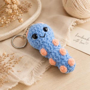 May include: A handmade blue crochet keychain shaped like a worm with peach-colored spots. The keychain has a smiling face with black eyes and a silver keyring attached. The background includes a neutral-colored fabric and a tag that says "handmade with love."