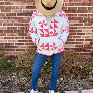 Puede incluir: Una persona con un sombrero de paja y una chaqueta acolchada blanca con un patrón geométrico rosa, vaqueros azules y zapatillas grises. La chaqueta tiene un bolsillo delantero y un cuello con cordón. El fondo es una pared de ladrillos y vegetación.
