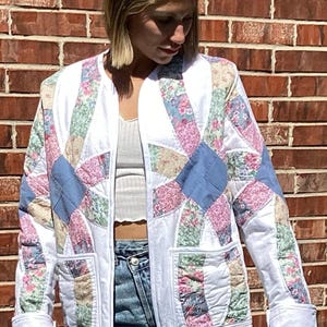 May include: A long-sleeve quilted jacket featuring a white base with a patchwork pattern of floral prints in pink, green, and blue, alongside solid blue fabric segments. The jacket has a collarless neckline, two front pockets, and white trim.